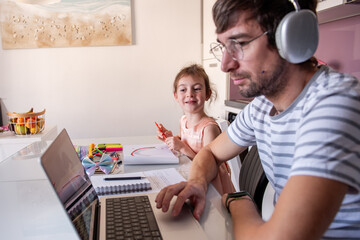 Father working from home, wearing noise-cancelling headphones, while little daughter happily drawing at the kitchen table, showcasing a loving family moment and modern parenting