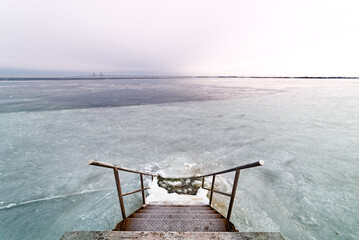 A stairway descends into a vast frozen Baltic sea under a grey sky, evoking isolation and...