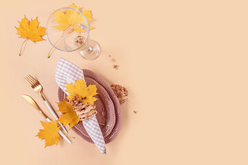 Autumn table setting on beige background. Oval plates, cutlery, glass, checkered napkin and dry yellow leaves. Autumn holiday mood, Halloween, Thanksgiving concept. Top view, copy space