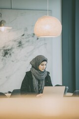 Inside a bright office with clean, minimalistic decor A worker in fusion attire, a traditional-style scarf paired with a modern jacket, types on their laptop The mood is calm and productive