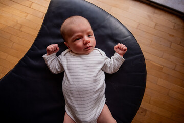 Newborn baby wearing a cozy white and gray striped bodysuit lies peacefully on a soft black leather cushion, raising tiny fists, captured from an overhead view