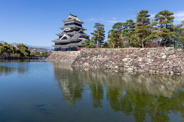 Matsumoto Castle stands above the still moat surrounded by stone walls pine trees and distant...