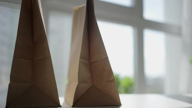 Brown paper bags resting on white table near window, carefully placing and arranging additional bag beside existing packaging, showcasing sustainable grocery and delivery container design