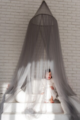 Young child sits contemplatively inside a mosquito net draped over a small set of stairs in a...