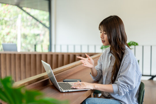 Woman explaining to online class or a meeting on laptop sitting at wooden counter in cafe or library - Powered by Adobe