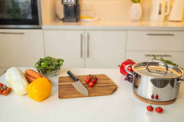 Fresh cherry tomatoes are sliced on a wooden cutting board in a bright kitchen. A sharp knife is used, and colorful vegetables like carrots, bell pepper, and spinach are nearby.