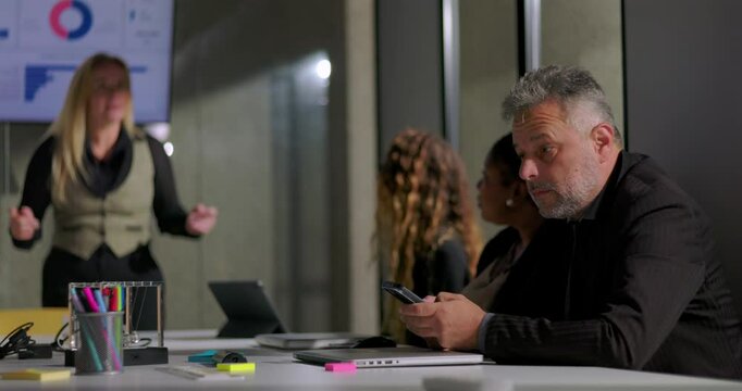Businessman discreetly checking phone during office meeting as female colleague presents, casual modern workplace dynamics with multitasking and quiet team engagement
