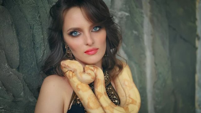 fantasy woman queen of snakes. Girl vampire fashion model posing with yellow tiger python snake animal. Beauty face lips blue eyes looking at camera. Lady love pet anaconda crawls over neck sexy body