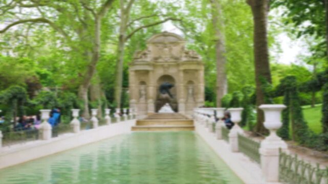 General view of the Medici Fountain and tourists near it surrounded by green trees in the Luxembourg Gardens on a sunny spring day. 6th arrondissement, Paris. Out of focus. Paris, France.