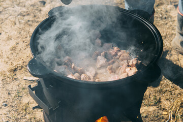 Pieces of meat frying in oil in a large cast iron cauldron with steam. Cooking outdoor in a big pot...
