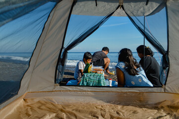 A family of four seen from inside a tent after completing their beachside meal, relaxing near the...
