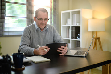 Senior man in sweater and glasses working from home using digital tablet