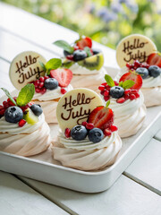Small pavlova mini cakes adorned with fresh berries like blueberries, strawberries, pomegranate seeds, kiwi slices, mint leaves and a white chocolate tag with text "Hello July".