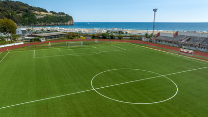 Aerial closeup of the midfield circle in an empty synthetic grass football field. In the background is the soccer goal and the sea.