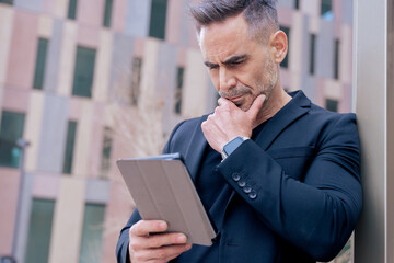 Caucasian mature man leaning on pole with a thoughtful expression while using a tablet. Dressed in black and surrounded by modern buildings, focused on work or information in urban setting