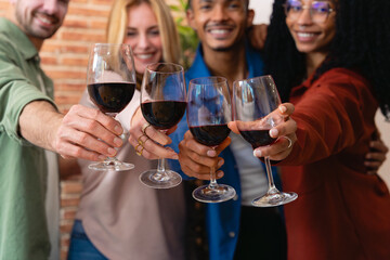 Smiling multiethnic friends raise glasses of red wine in a toast. Indoor setting with exposed brick wall, joyful atmosphere, casual outfits, and strong sense of celebration and friendship. 