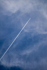 An airplane contrail streaks across a blue sky with wispy clouds.