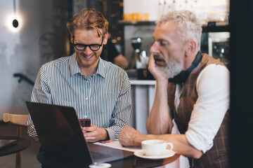 Red-haired businessman with laptop sits on bleachers discussing work with older gray-bearded colleague holding papers. Office has natural light, casual seating, relaxed startup environment