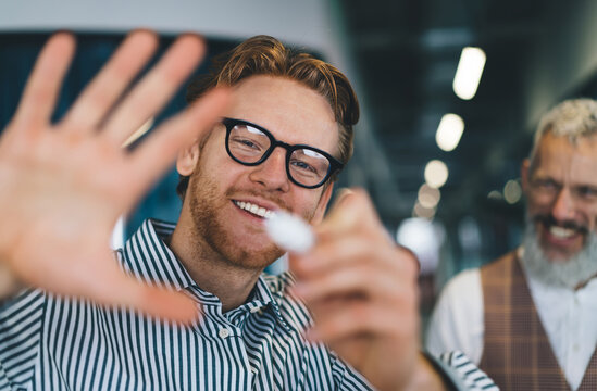 Cheerful young Caucasian man with red hair and glasses playfully poses for the camera, smiling and raising hand forward in modern office interior, middle-aged man blurred in the background