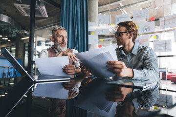 Two businessmen, one mature and one younger, standing near a glass board filled with business...