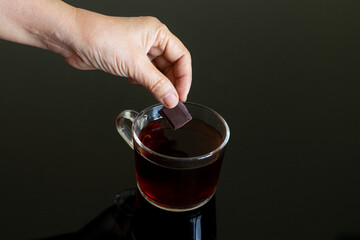 Cacao mass with black coffee, A hand pick up a piece of cacao mass or chocolate liquor to hot black coffee in a clear glass on black glass table with reflection close up
