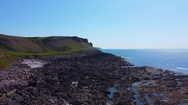 Slow Rising Drone Aerial View of Steep Cliff Tops with Bay at Low Tide and Sun Reflecting on Sea Water Creating Wide Picturesque View.