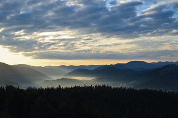 Sun rising over the Carpathian mountains, panoramic view. Silhouettes of mountain peaks. Morning fog. Dense forest in the foreground.