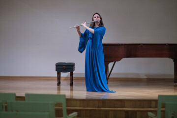 Woman in flowing blue dress performs on stage, elegantly playing the flute. Emotionally engaged, she stands before an empty hall, capturing the essence of music expression.