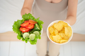 Pregnant woman holding both wholesome salad and salty chips. Authentic snapshot of dietary...