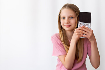 Fototapeta premium Girl smiling while holding chocolate bar. Young girl in pink shirt smiles softly while presenting a large dark chocolate bar wrapped in foil against a white background. Space for text.