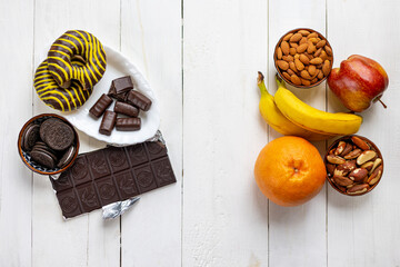Healthy vs Unhealthy Food Choices Comparison. Top view of healthy fruits and nuts contrasted with sugary chocolate, cookies, and donuts on a white wooden background.