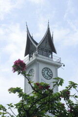 Fototapeta premium A close-up of the iconic Jam Gadang clock tower in Bukittinggi, Indonesia, featuring traditional Minangkabau architecture. Pink flowers in the foreground add a vibrant touch to the elegant structure u