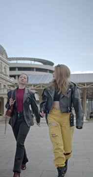 Two young women, dressed in stylish outfits and radiating joy, stroll through a city square, holding hands and sharing laughter.