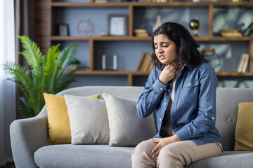 Fototapeta premium A young woman experiences throat pain while sitting on a comfortable sofa in her living room at home.