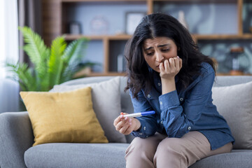 A worried woman is looking at a pregnancy test while sitting on a couch in her home.