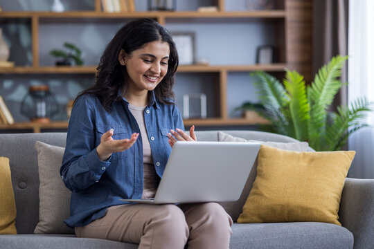 A cheerful woman has a video call on her laptop while sitting on a comfortable sofa at home.