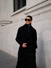 Portrait of handsome Chinese young man wearing black overcoat posing in the street, young guy with black short hair all in black with urban background. Male fashion, cool Asian young man lifestyle.