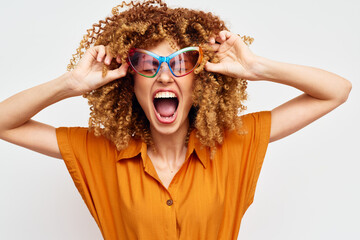 Happy woman with curly hair wearing colorful sunglasses, expressing joy against a light background,...