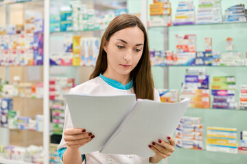Young adult female pharmacist with long brown hair reviews documents in pharmacy. Caucasian woman...