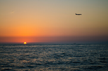 Airplane glides above calm ocean, silhouetted against vibrant sunset. Orange and purple tones...