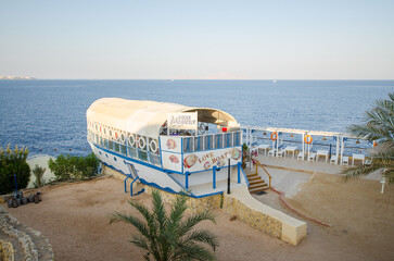 Resort with boat-shaped restaurant offering stunning ocean views. Palm trees frame sandy foreground. Soft, natural lighting enhances tranquil ambiance