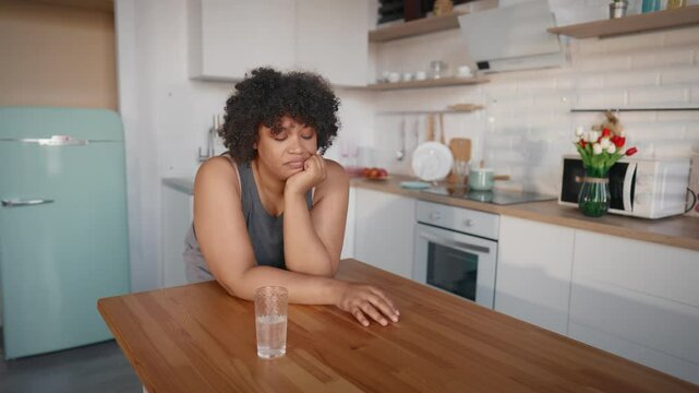 Low calories diet for lose weight, sad african american woman drinking water. Medium portrait of unhappy overweight lady in home kitchen, feel hungry, want to eat, hormonal mood swings in pregnancy