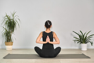 Young woman with medium skin tone sits cross-legged on yoga mat, performing reverse prayer pose....