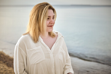 Young woman in white blouse stands on tranquil beach, gazing thoughtfully at calm sea, evoking...