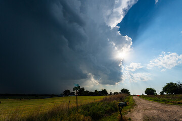 Sun rays shine through storm clouds over a quiet country road, creating a dramatic sky with rich texture, shadow, and moody atmosphere, blending beauty in nature with the power of weather.