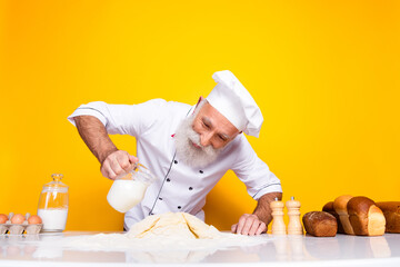 Senior chef in a white uniform preparing dough in a bright kitchen with yellow background,...