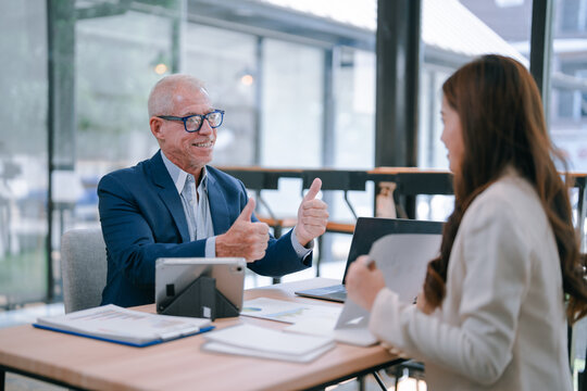 Senior business manager showing thumbs up gesture of approval and appreciation to his young female colleague during a business meeting in a modern office