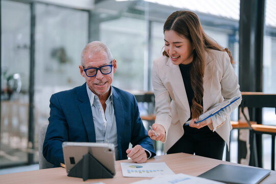 Two smiling business colleagues are collaborating on a project, analyzing financial charts and data displayed on a digital tablet and printed documents, working together at an office desk