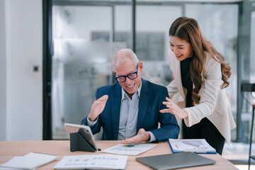 Senior manager and his young assistant are smiling and looking at financial charts on the table while having a video conference with a tablet in the office