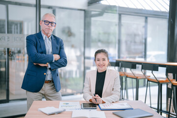 Senior manager standing with arms crossed and young businesswoman sitting at desk working with...